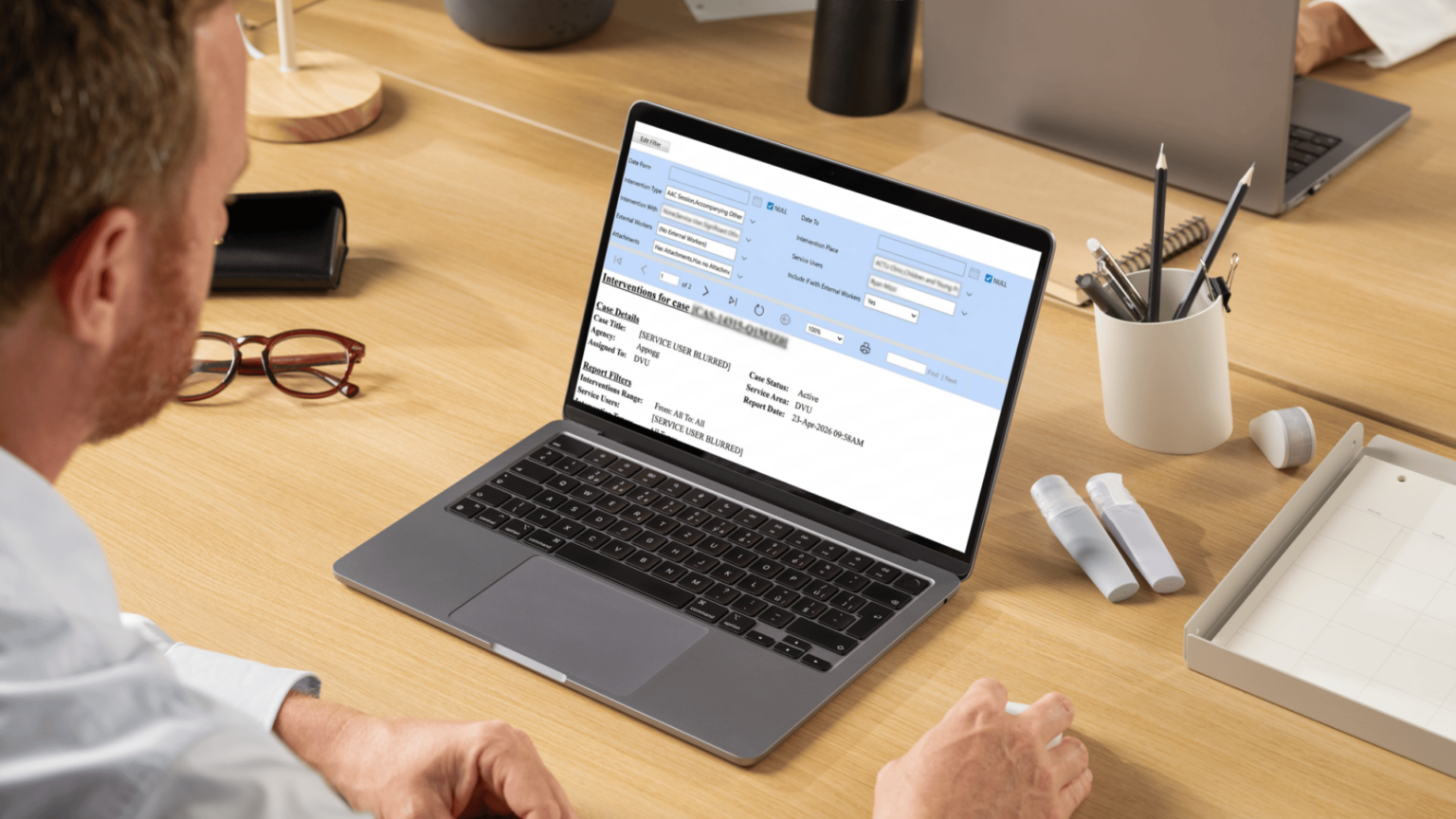 Person at desk viewing a Microsoft Dynamics 365 social welfare case management screen on a laptop, showing an “Interventions for Case” report with case details, service area and reporting filters